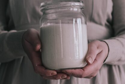 A woman, dressed in a beige apron, holding a glass jar filled with sourdough starter