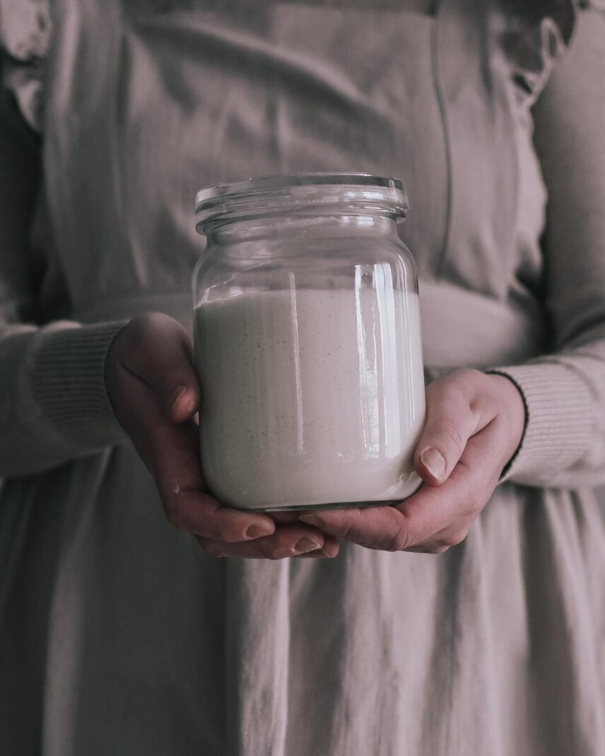 A woman, dressed in a beige apron, holding a glass jar filled with sourdough starter