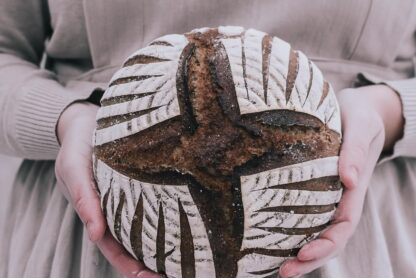 woman, dressed in a beige apron, holding a rye sourdough bread