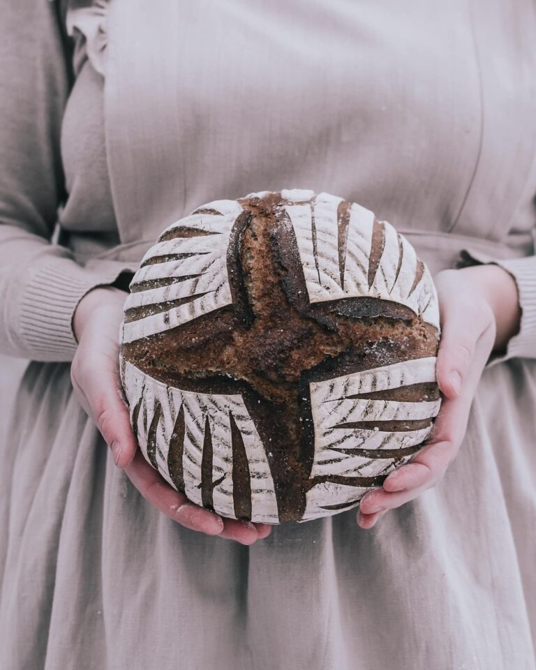 woman, dressed in a beige apron, holding a rye sourdough bread